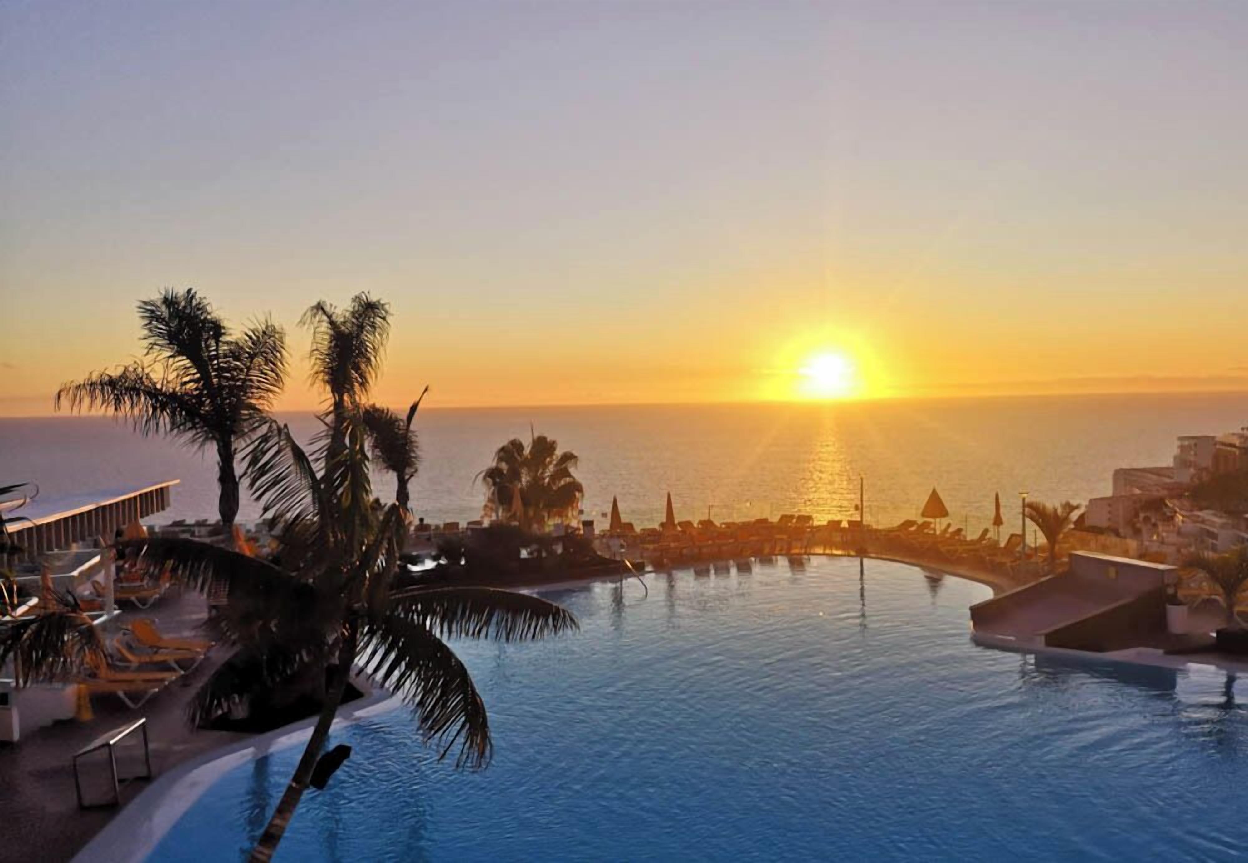 Piscina de un resort con palmeras y vista al atardecer.