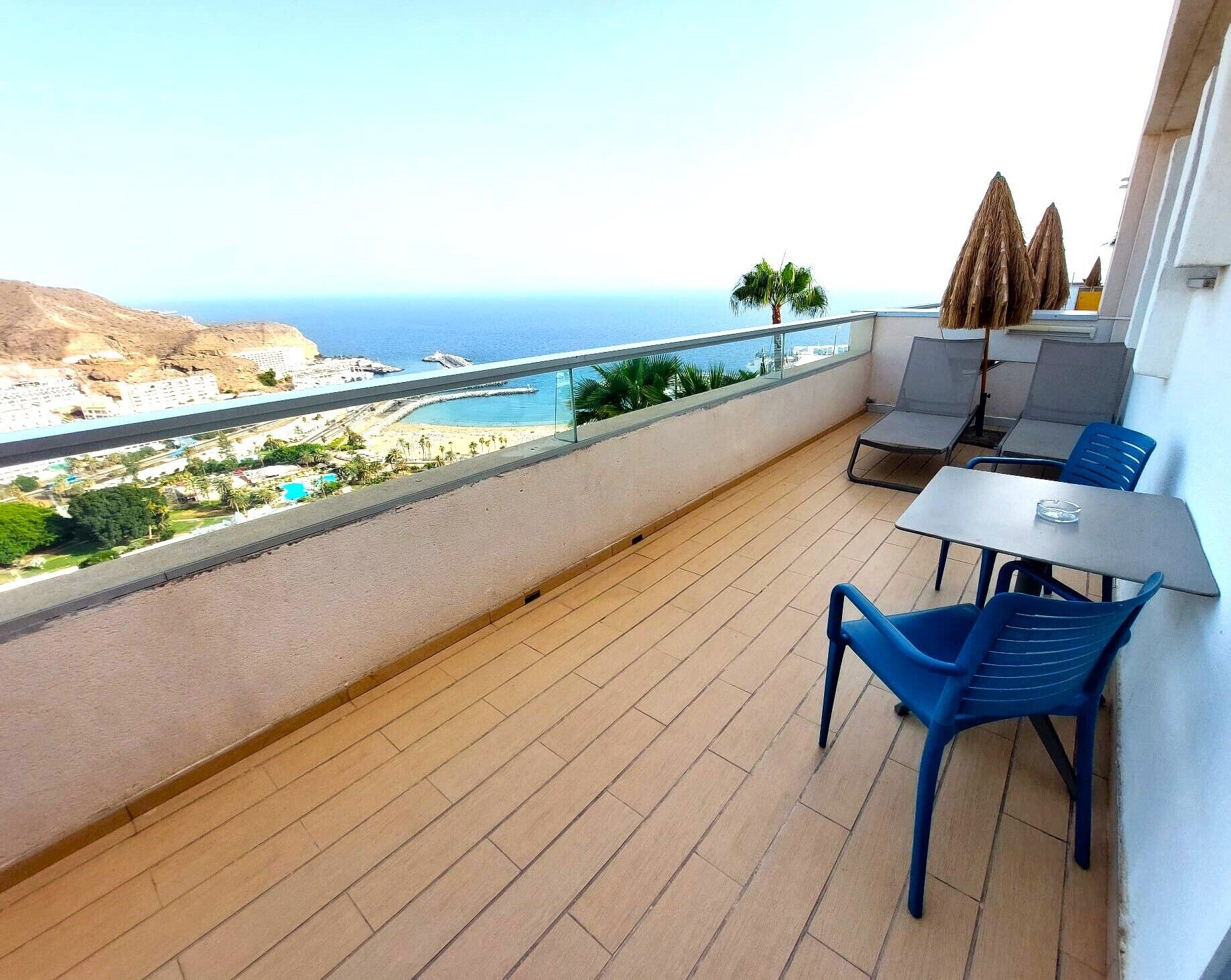 Een balkon met uitzicht op zee en een strand. Er zijn twee blauwe stoelen en een tafel.