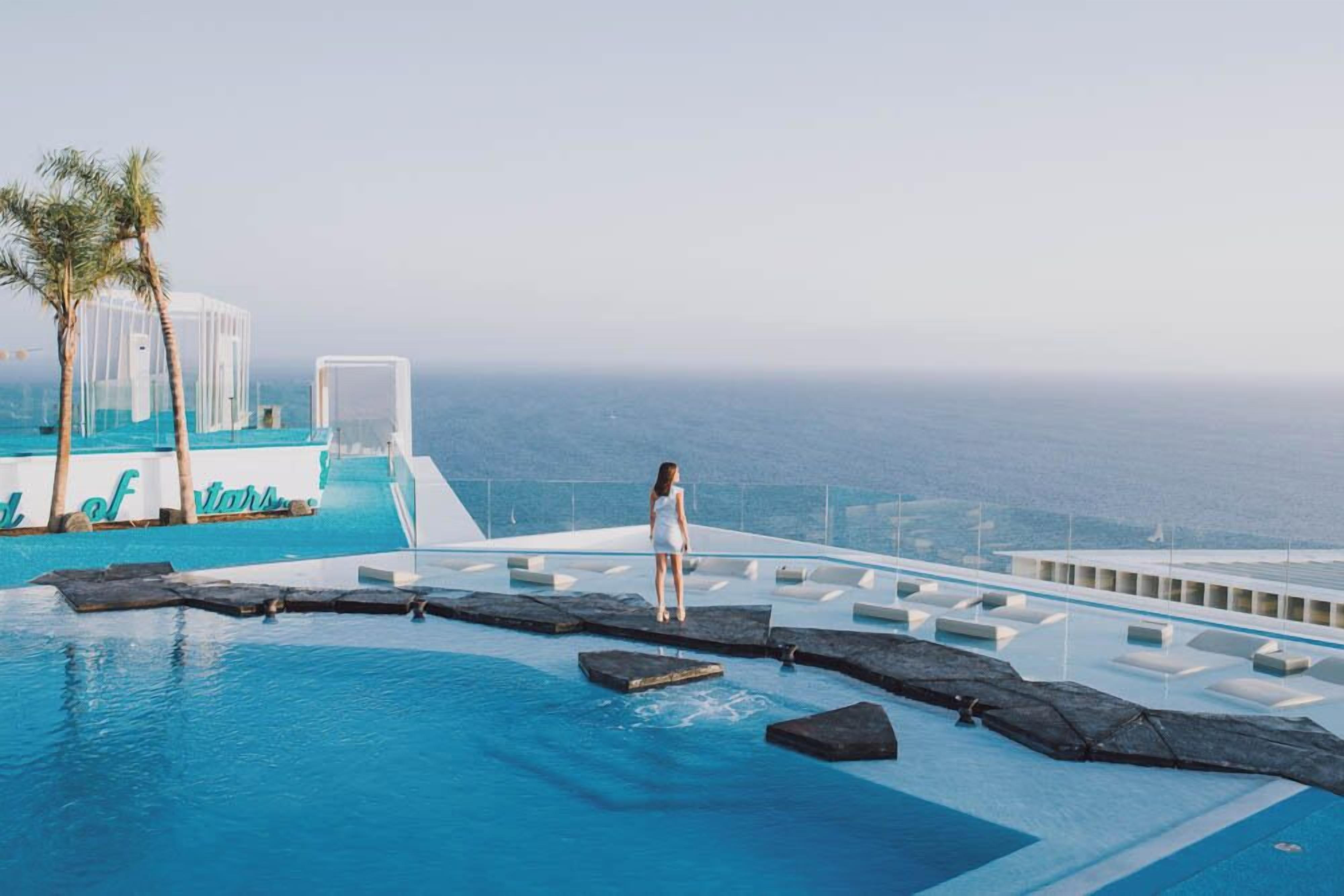 A pool area with a person standing on a platform, palm trees, and a view of the ocean.