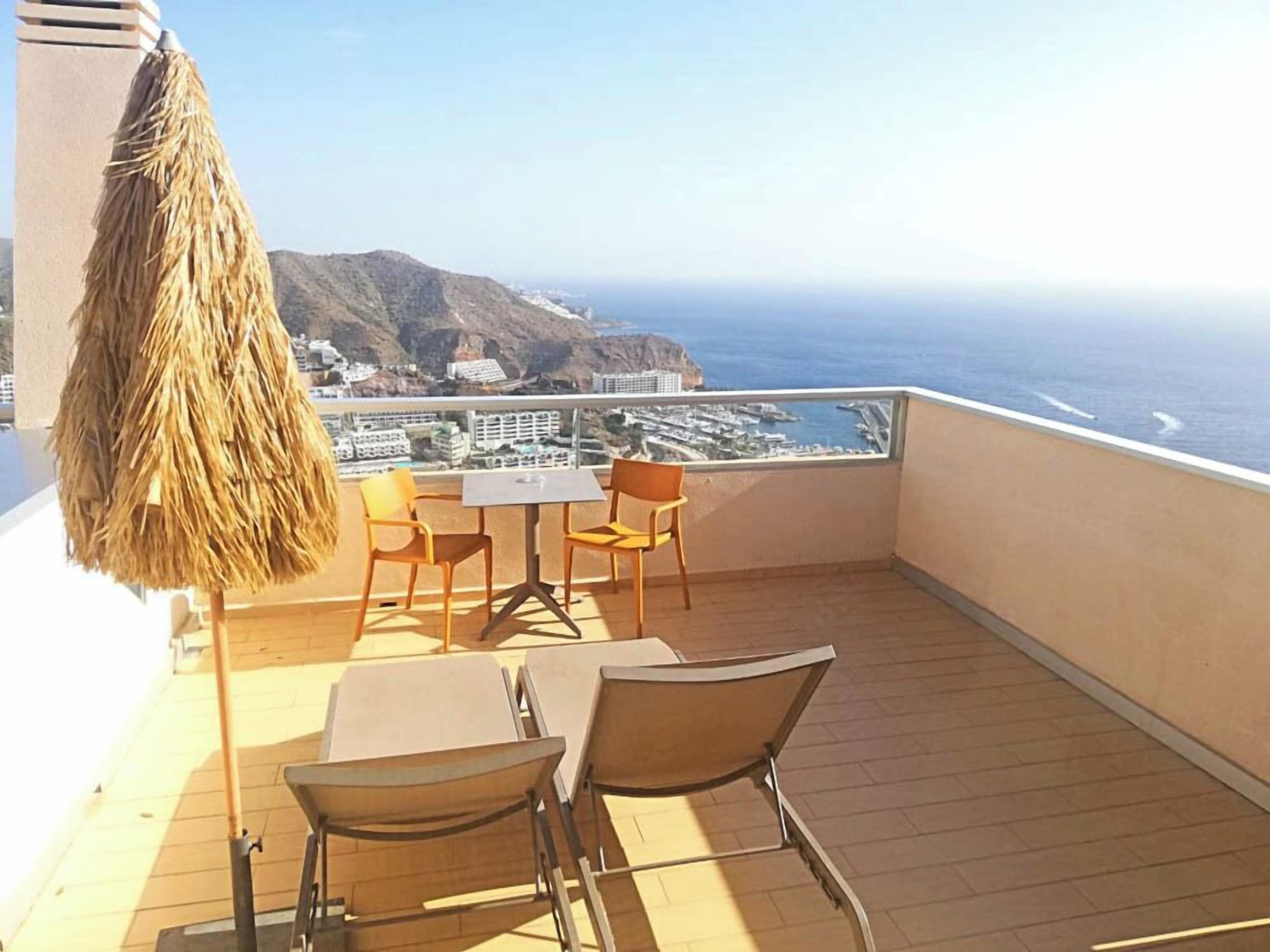 A balcony with wooden flooring, a table with chairs, and a thatched umbrella.