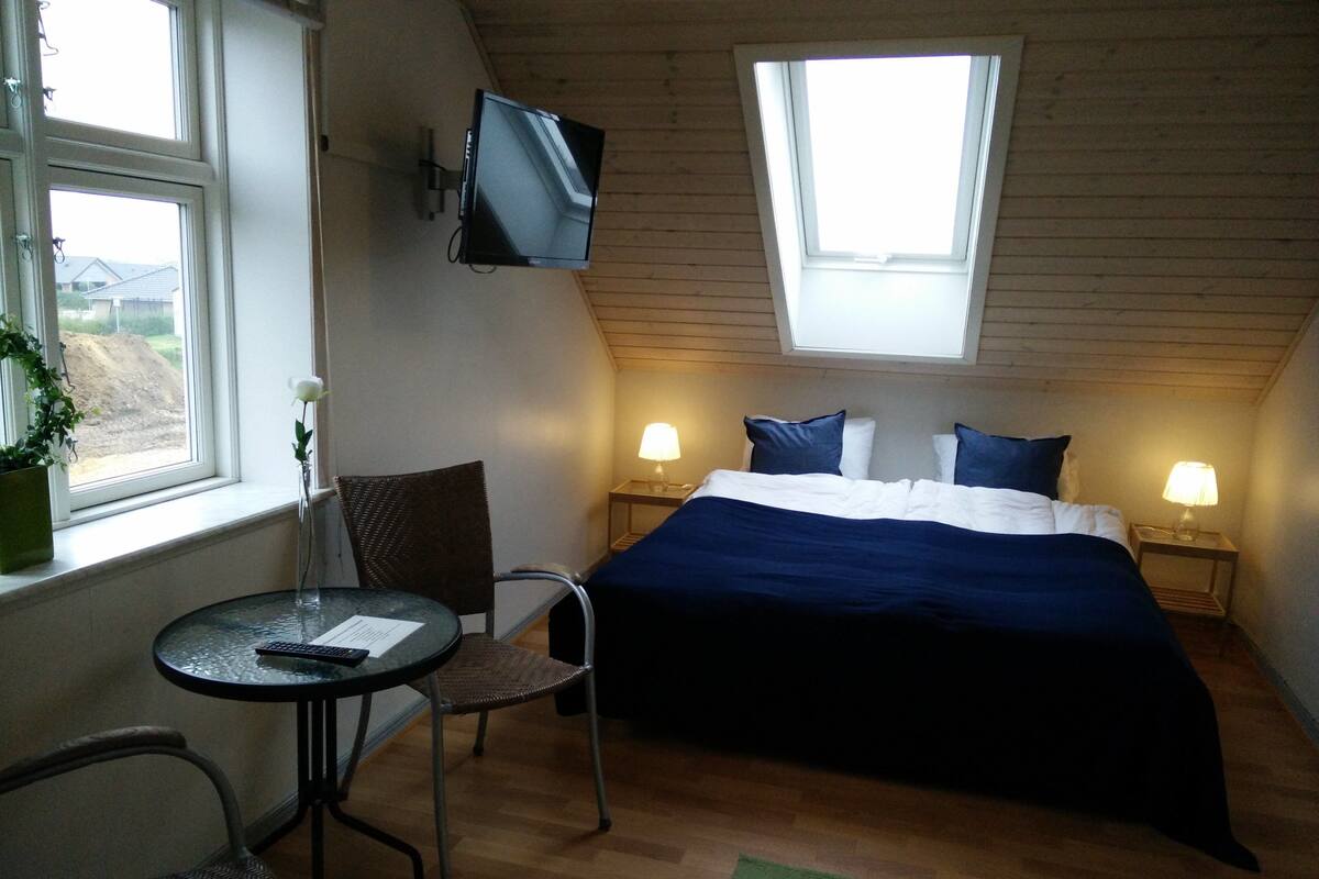 A bedroom with a bed, two bedside lamps, a TV mounted on the wall, a chair, a small table, and a window with a view of the outside.