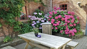 Ein hölzerner Picknicktisch mit Bänken, ein Blumentopf und eine Holzhütte in einem Garten mit Ziegelmauern und blühenden Pflanzen.