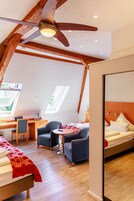 Un dormitorio con techo inclinado, vigas de madera, una cama con colcha roja y blanca, dos sillas, un escritorio con ordenador y una ventana con vistas a la vegetación.