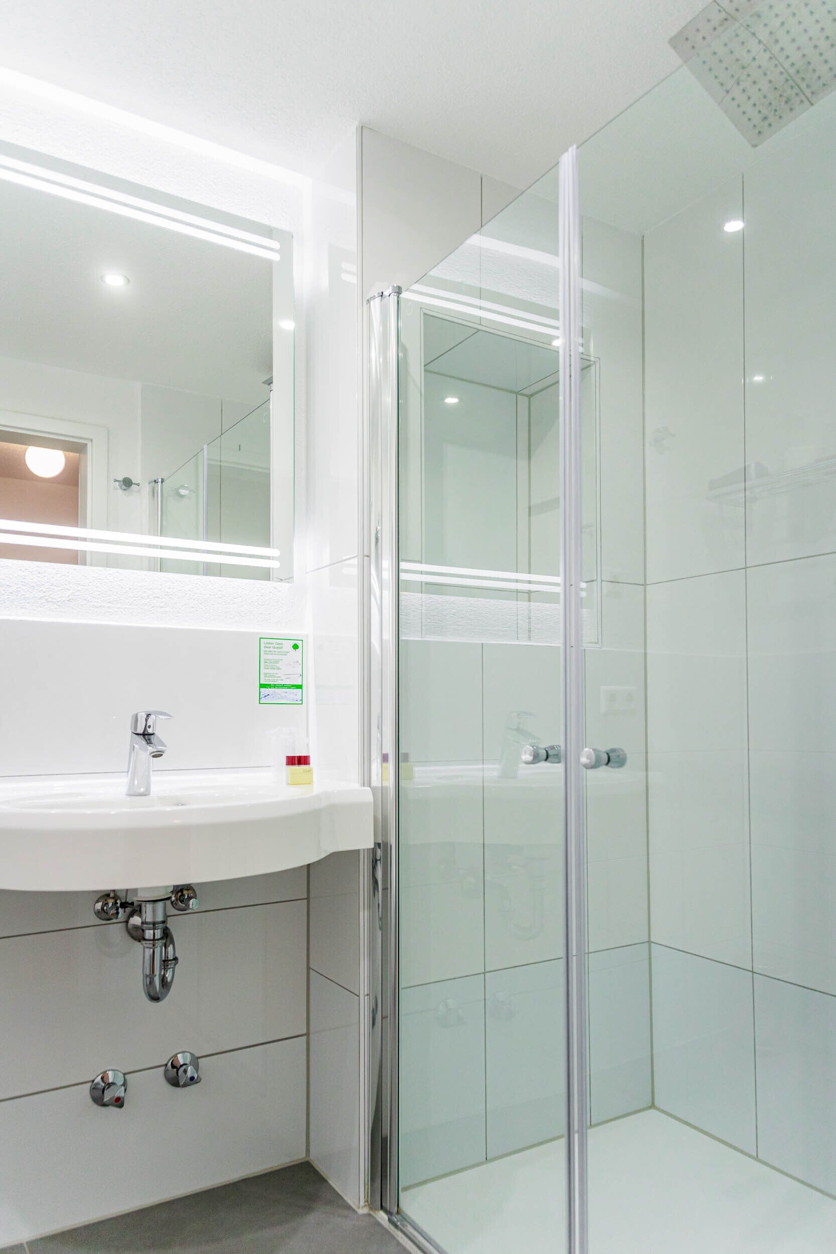 Une salle de bain moderne avec une douche à l’italienne en verre, un lavabo blanc avec un miroir au-dessus, et un chauffe-eau visible.