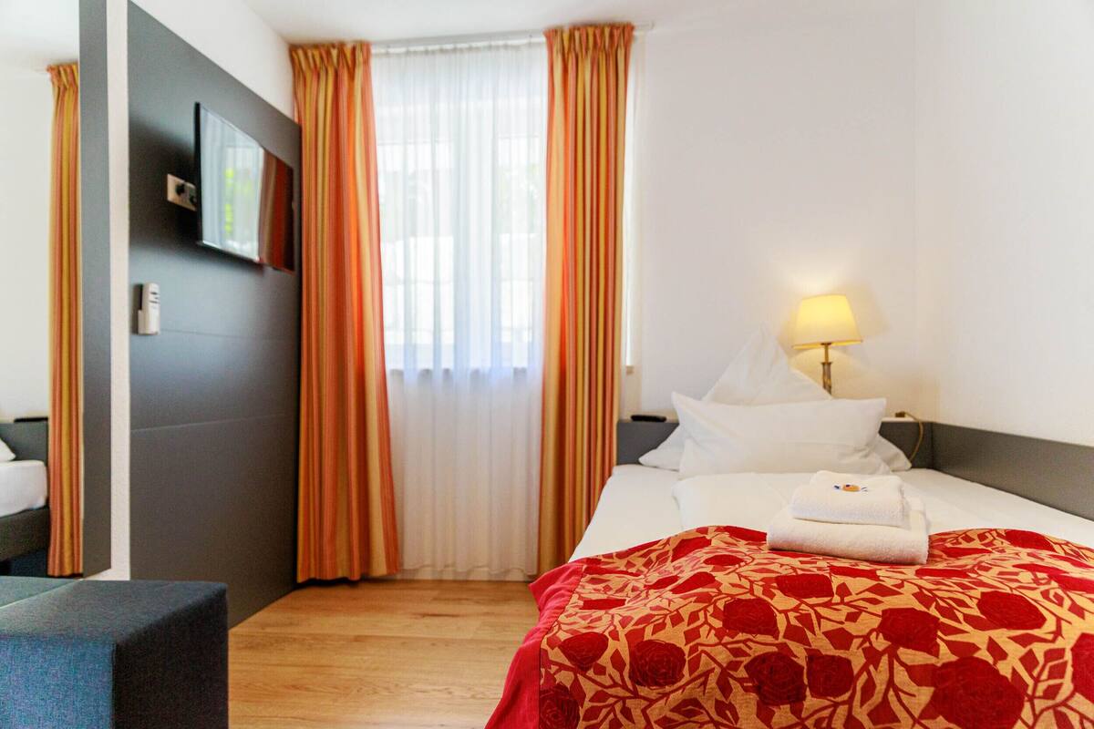Hotellihuone, jossa on sänky, yöpöytä, lamppu ja ikkuna, jossa on verhot.