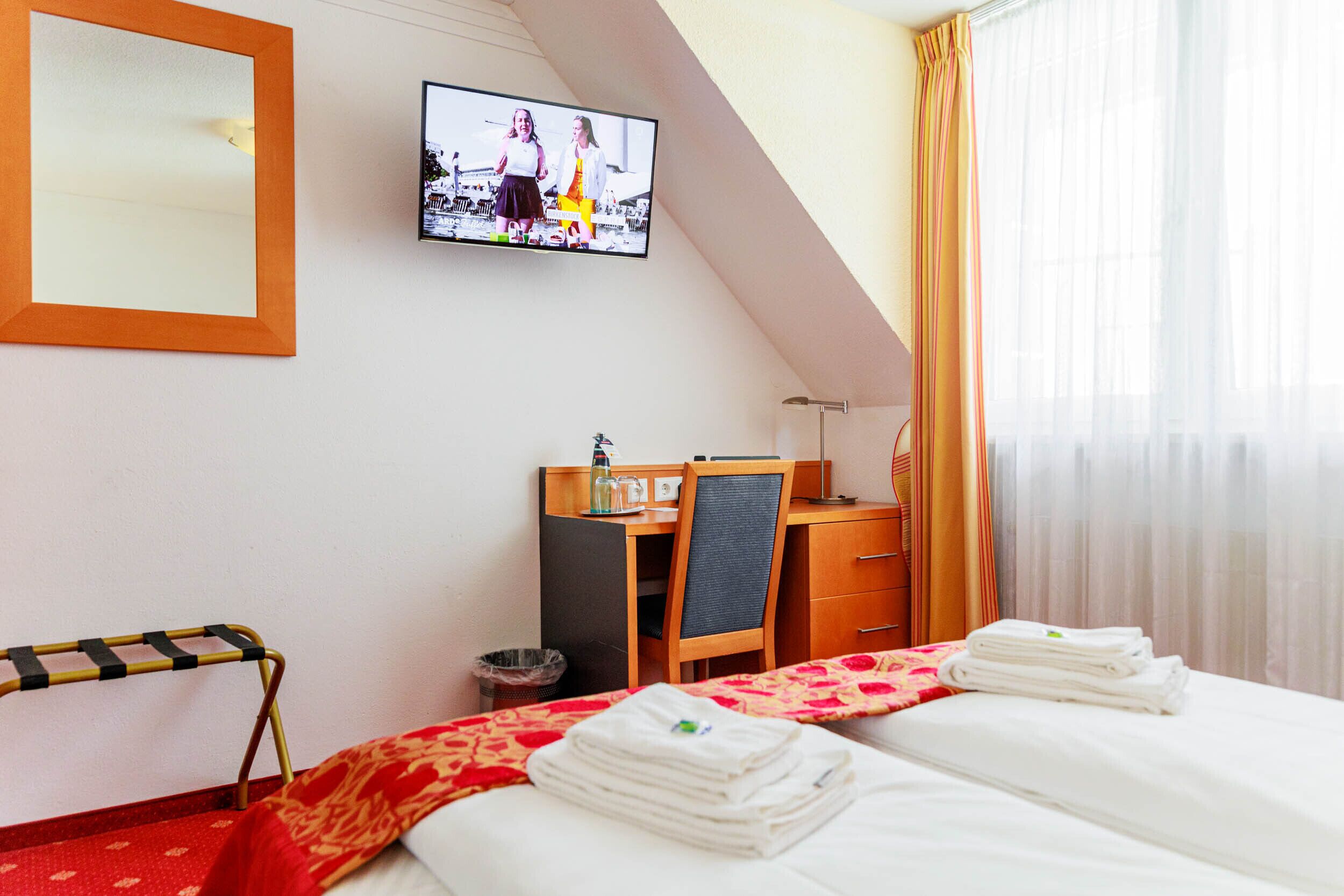Habitación de hotel con cama, escritorio, silla, televisor y ventana con cortinas.