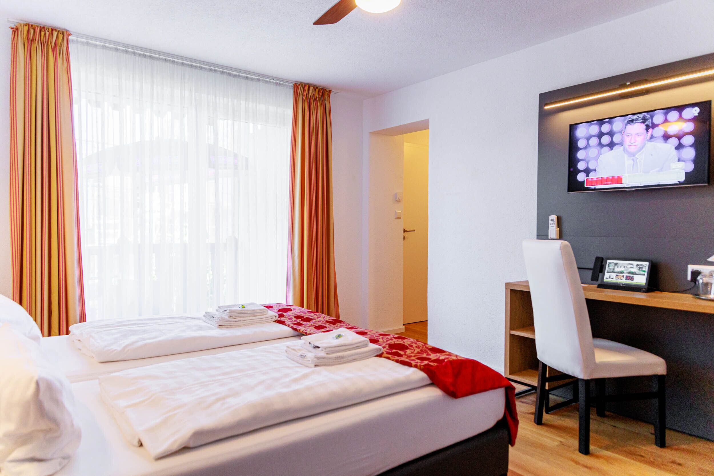 Habitación de hotel con dos camas, un televisor en la pared, un escritorio con una silla y una manta roja.