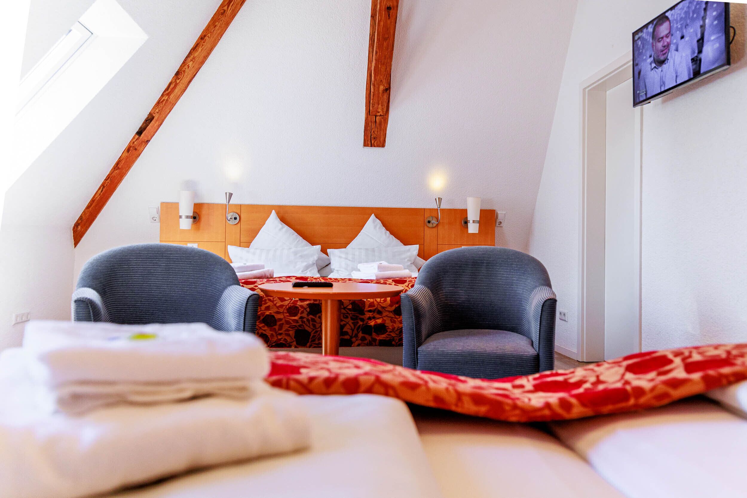A modern hotel room with a wooden ceiling, a bed with a red and white patterned blanket, two grey armchairs, a small round table with a remote control, and a television mounted on the wall.
