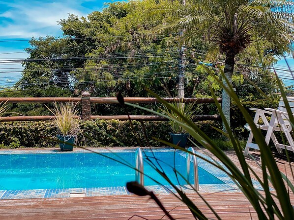 A swimming pool with a wooden deck, surrounded by lush greenery and trees. - Pousada Solar de Búzios (Búzios)