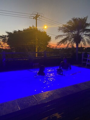 A swimming pool at sunset with two people in the water, surrounded by palm trees and a fence. - Pousada Solar de Búzios (Búzios)