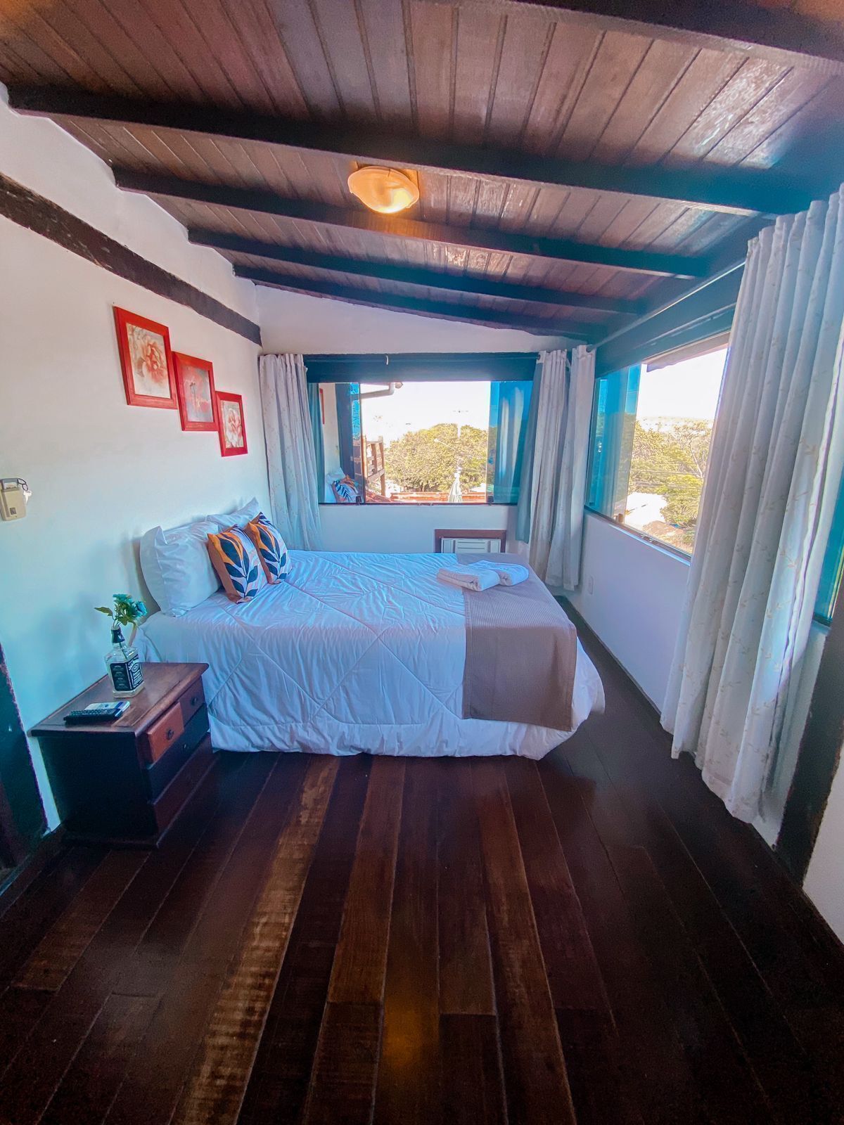A bedroom with a wooden ceiling, a bed with a white duvet, a nightstand with a bottle, and a window with curtains.