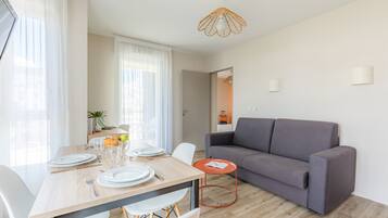 Una sala de estar moderna con un sofá gris, una mesa de comedor de madera dispuesta con platos y vasos, y una pequeña mesita auxiliar naranja.