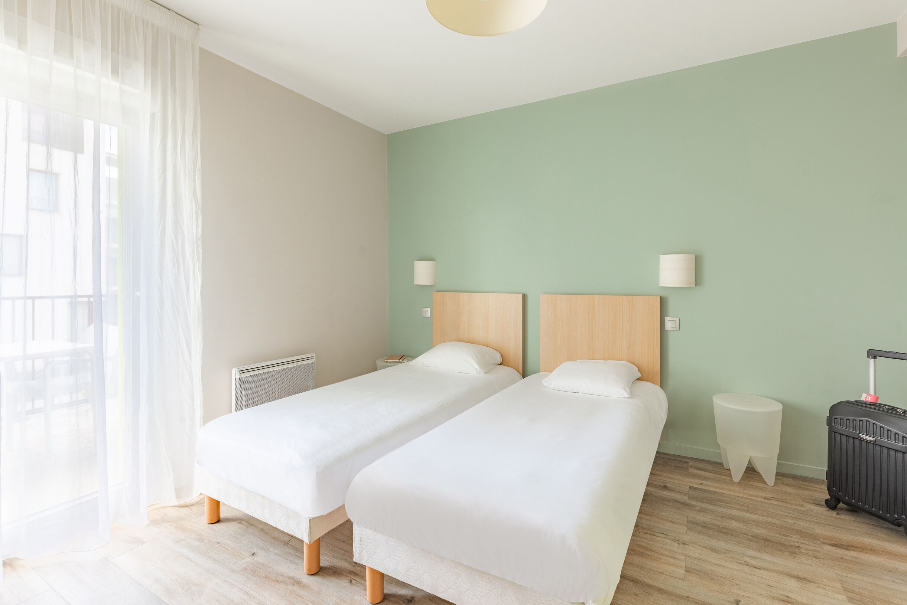 A hotel room with two single beds, a bedside table, a lamp, a radiator, and a suitcase.