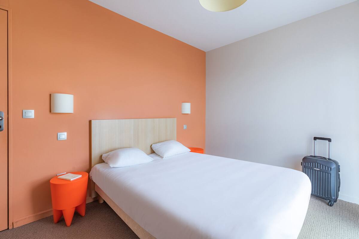 A hotel room with a large bed, an orange accent wall, a small orange side table, and a grey suitcase.