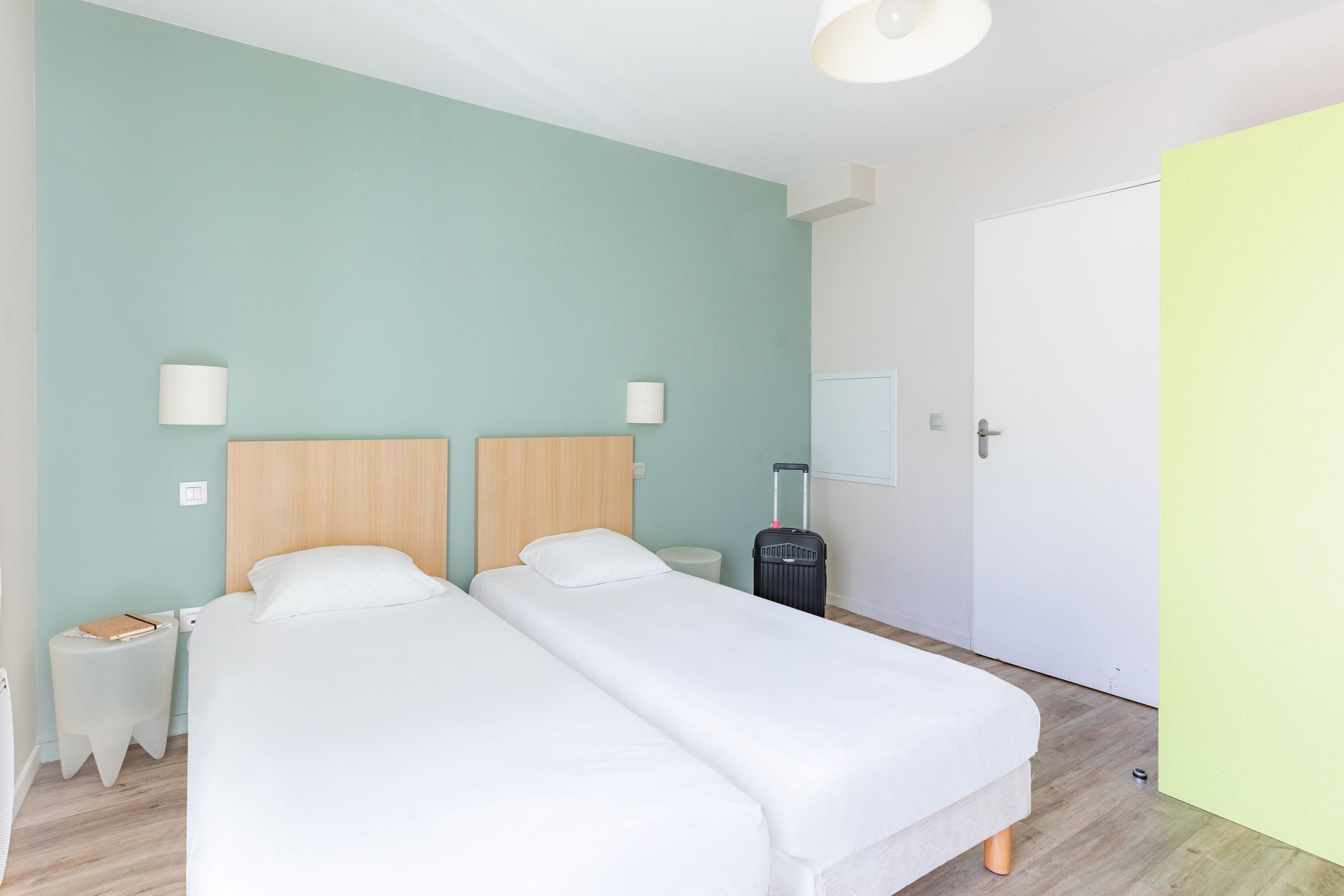 Une chambre d’hôtel avec deux lits, une table de chevet, une lampe, une valise et une armoire.