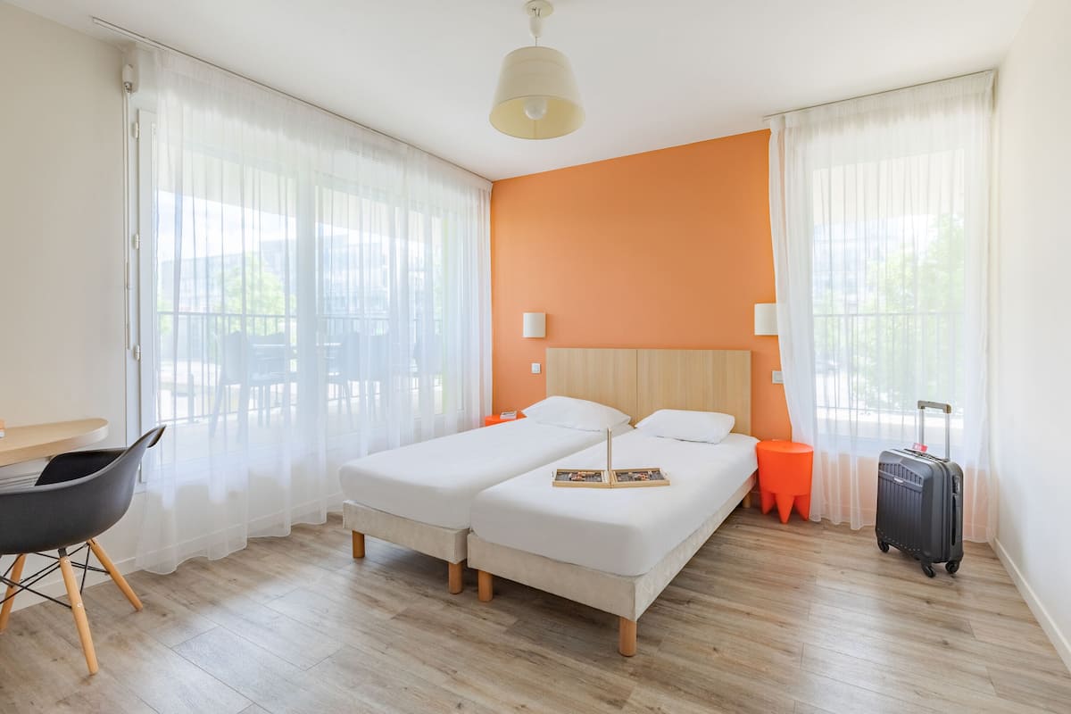A hotel room with two beds, a desk, a chair, a suitcase, and a balcony with a table and chairs.