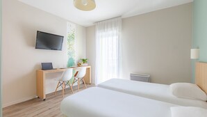 A modern hotel room with a wooden desk, a flat-screen TV, two beds, a window with sheer curtains, and a ceiling light. - Appart'City Confort Tours (Tours)