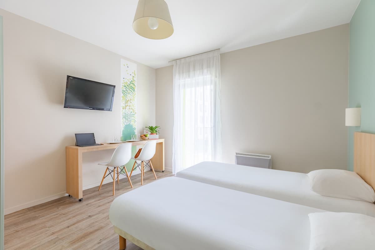 A modern hotel room with a wooden desk, a flat-screen TV, two beds, a window with sheer curtains, and a ceiling light.
