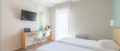 A modern hotel room with a wooden desk, a flat-screen TV, two beds, a window with sheer curtains, and a ceiling light.