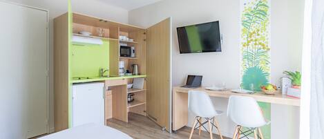 Quarto moderno e compacto com cozinha compacta, escrivaninha com computador, duas cadeiras e uma cama.