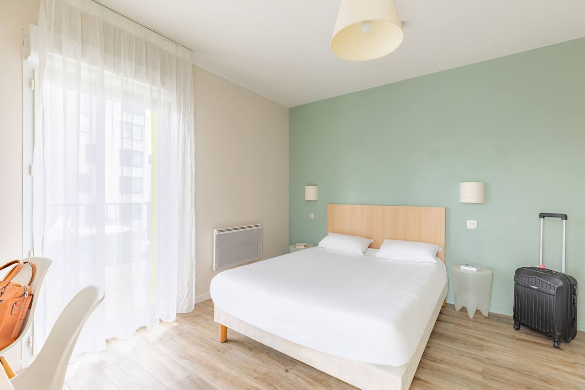 A hotel room with a bed, a chair, a suitcase, a nightstand, and a lamp.