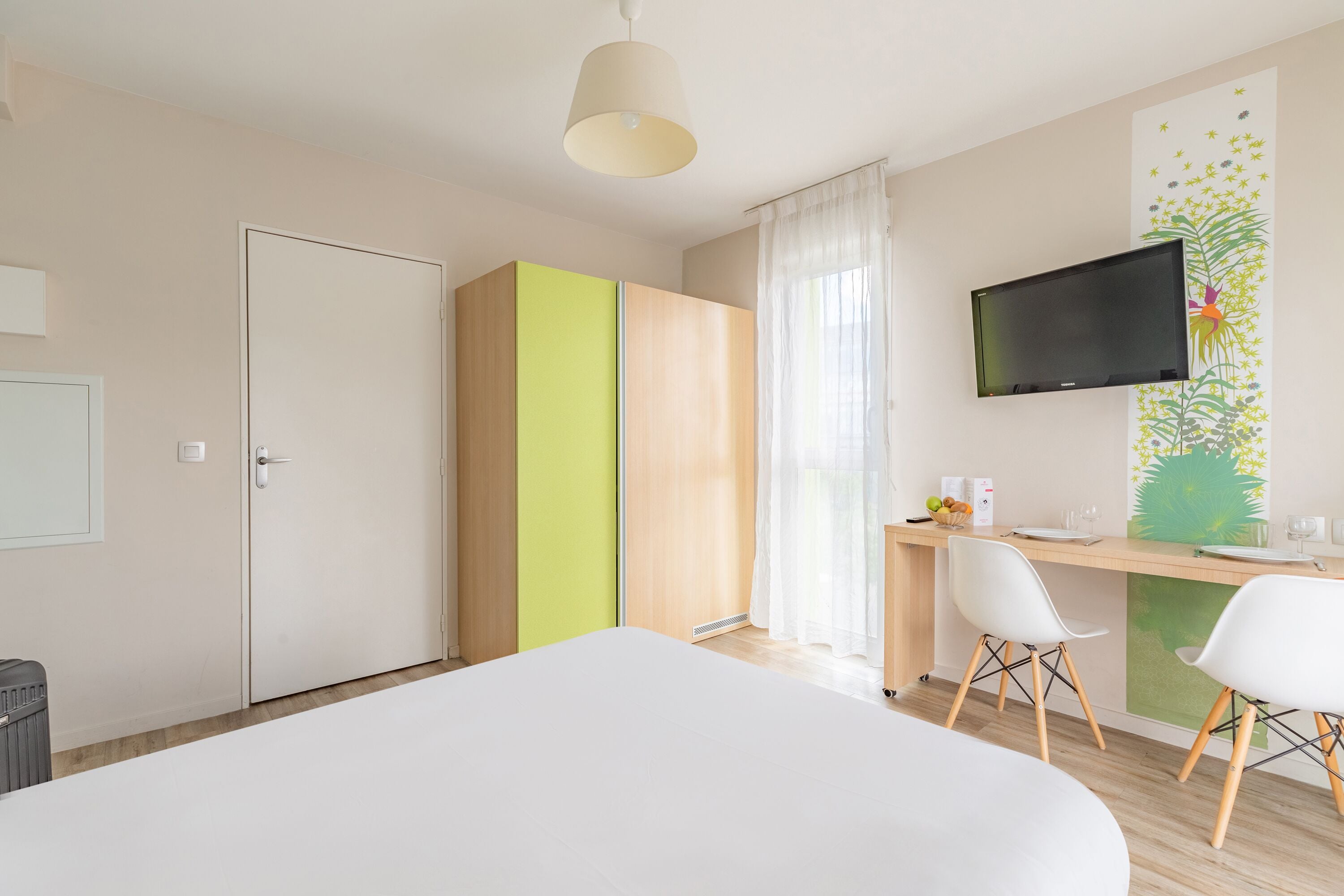 A modern hotel room with a bed, a desk with chairs, a television, and a wardrobe.