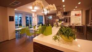A modern interior with a dining area, kitchen, and bar. There are green chairs, a white counter, and potted plants. - Hotel Casa Serena (Malcesine)