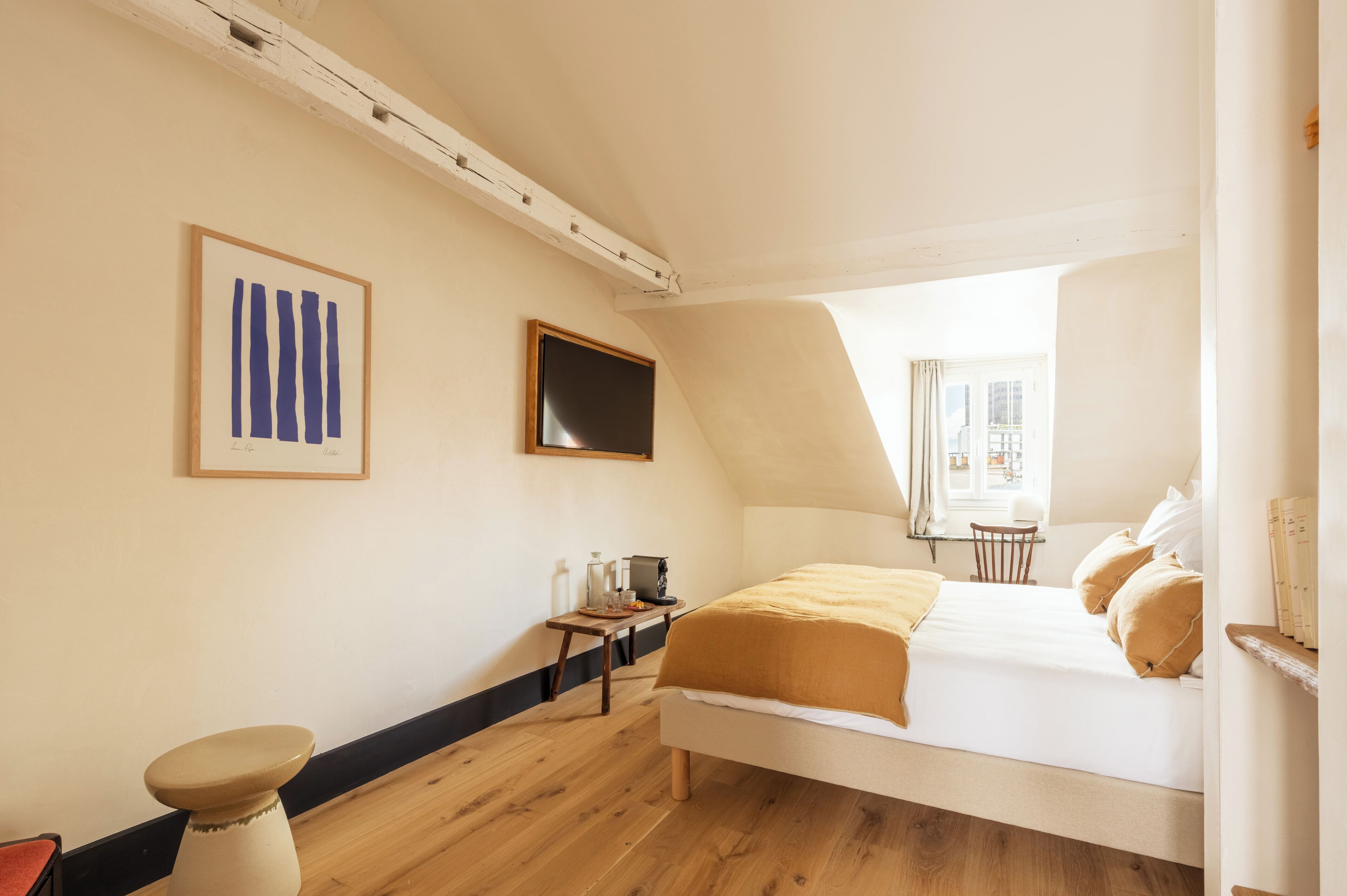 Un dormitorio con cama, escritorio, silla, taburete, un cuadro enmarcado y una ventana.