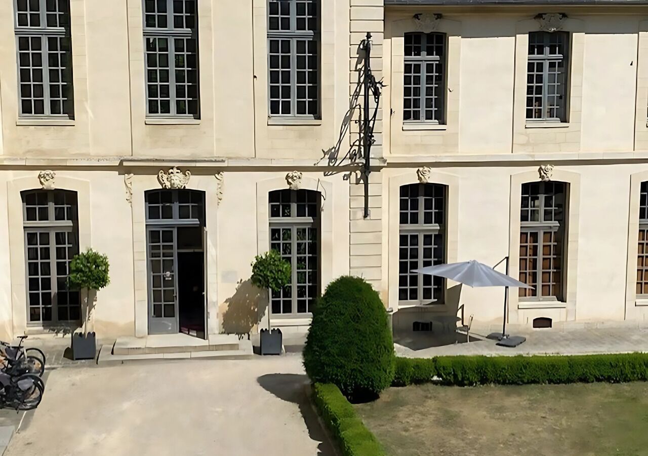 Un bâtiment doté de nombreuses fenêtres et portes, d’une cour pavée et de quelques plantes en pot.
