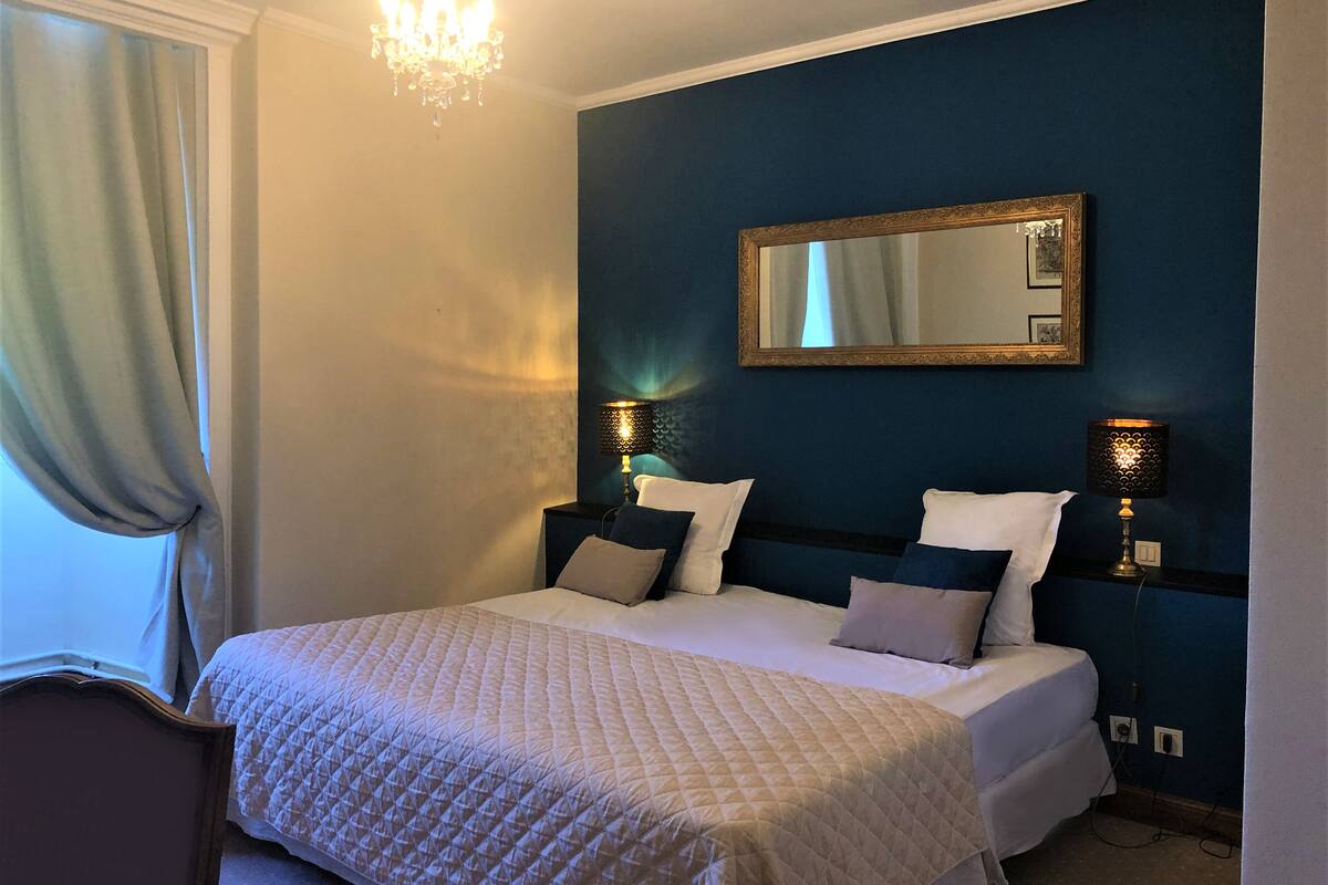 Un lit bien fait, recouvert d’une courtepointe, deux lampes de chevet, un miroir au mur et un lustre.