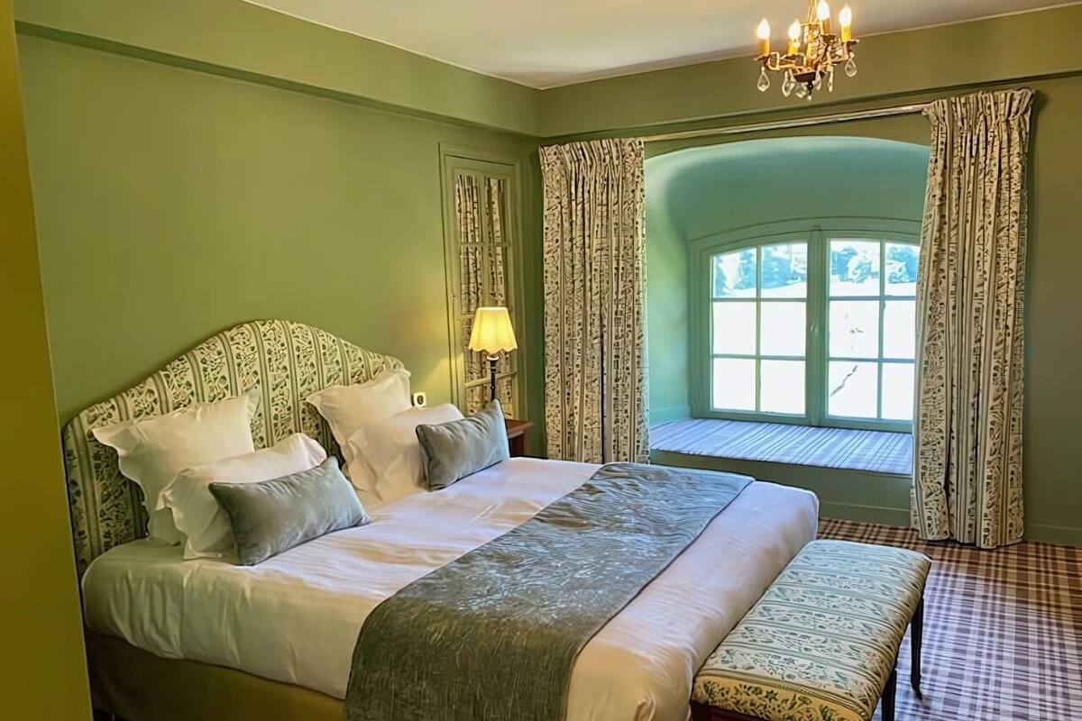 Un lit bien fait, avec des oreillers et un banc, une table de chevet avec une lampe, et une fenêtre avec des rideaux.