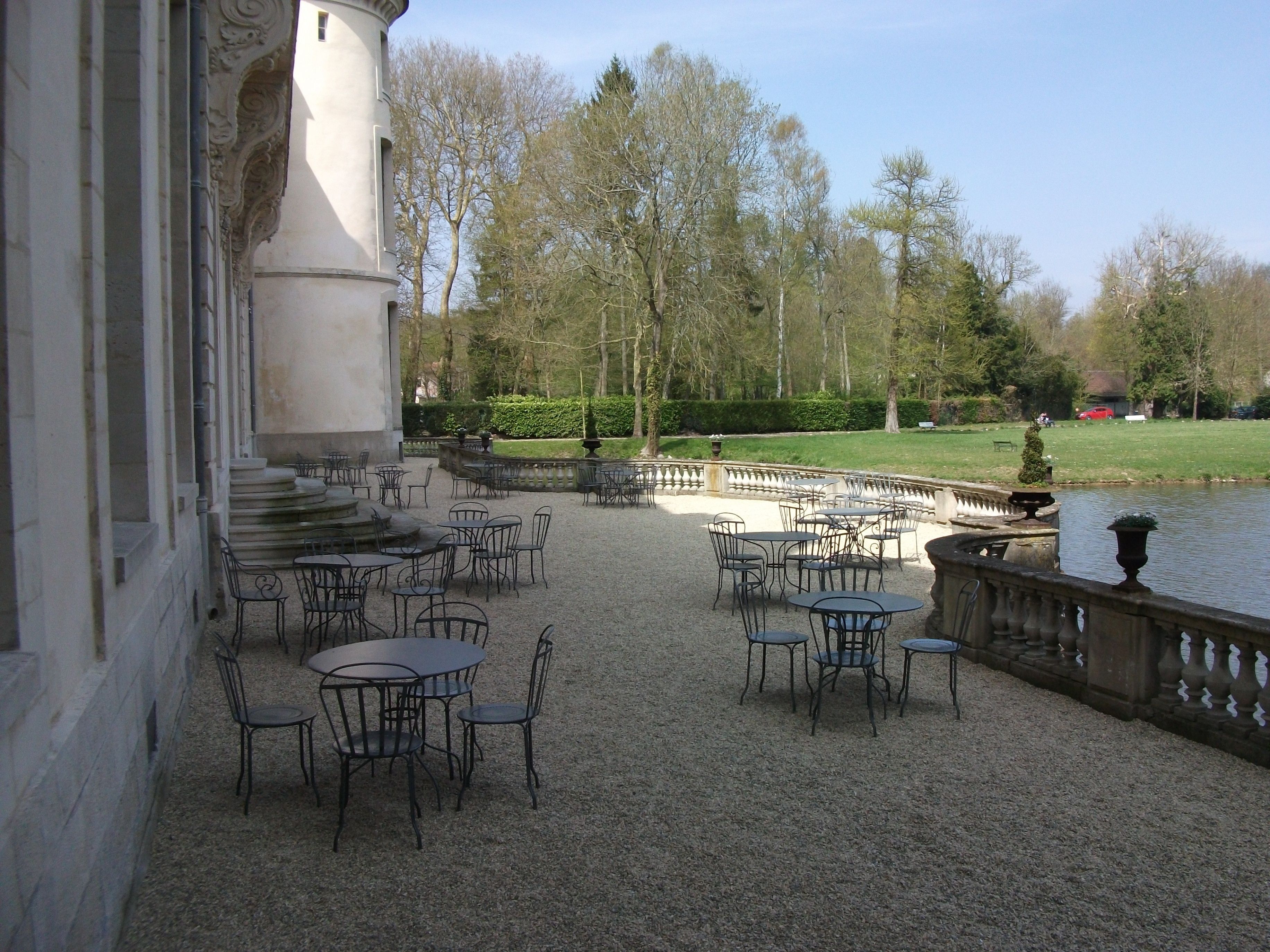Une terrasse extérieure avec des tables et des chaises, une balustrade en pierre et une étendue d’eau en arrière-plan.