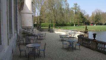Une terrasse extérieure avec des tables et des chaises, une balustrade en pierre et une étendue d’eau en arrière-plan.