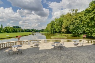 Un patio jardín con mesas y sillas con vistas a un lago, rodeado de exuberante vegetación y un cielo despejado.