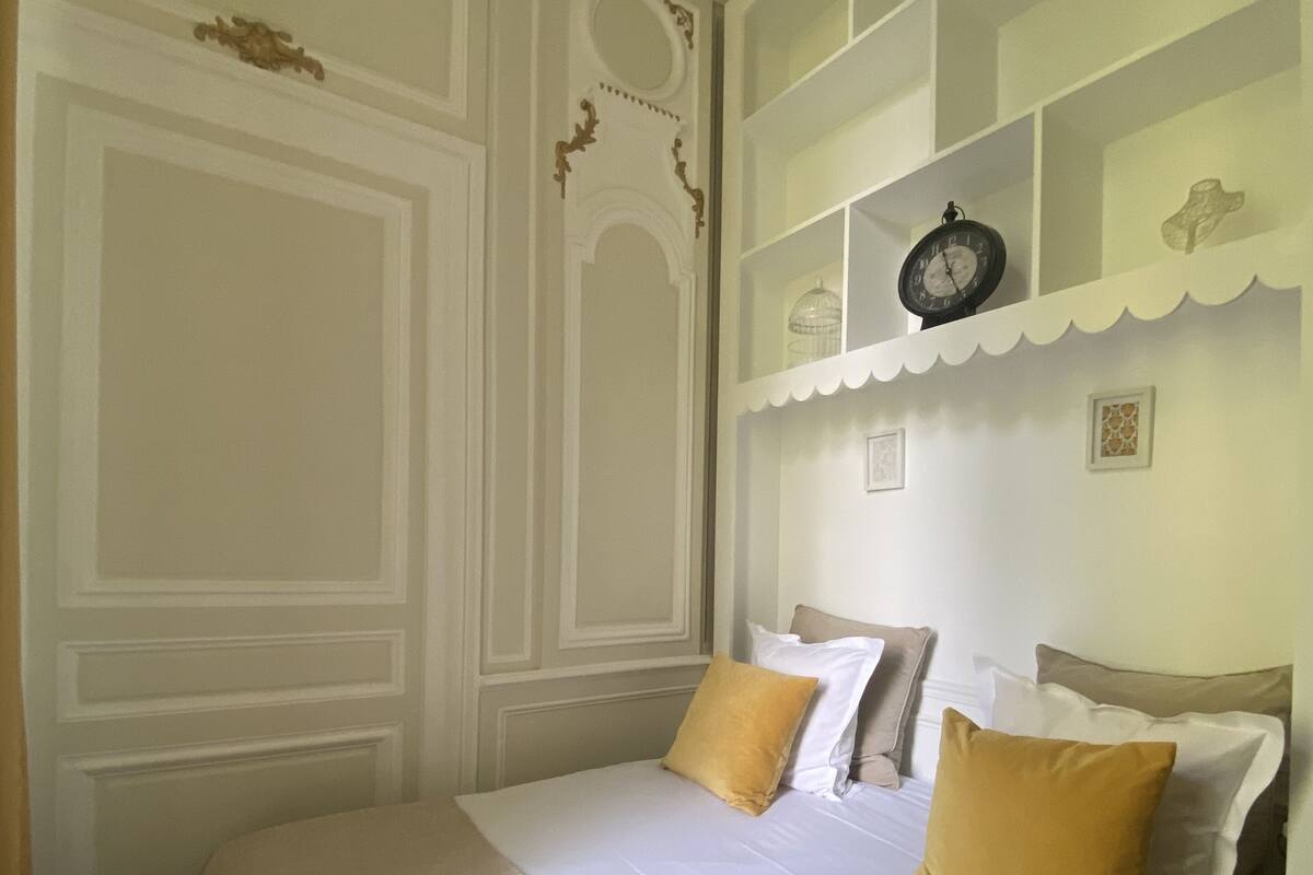 Un lit bien fait, avec des oreillers blancs et jaunes, une horloge au mur et une étagère décorative sur laquelle est posée une cage à oiseaux.