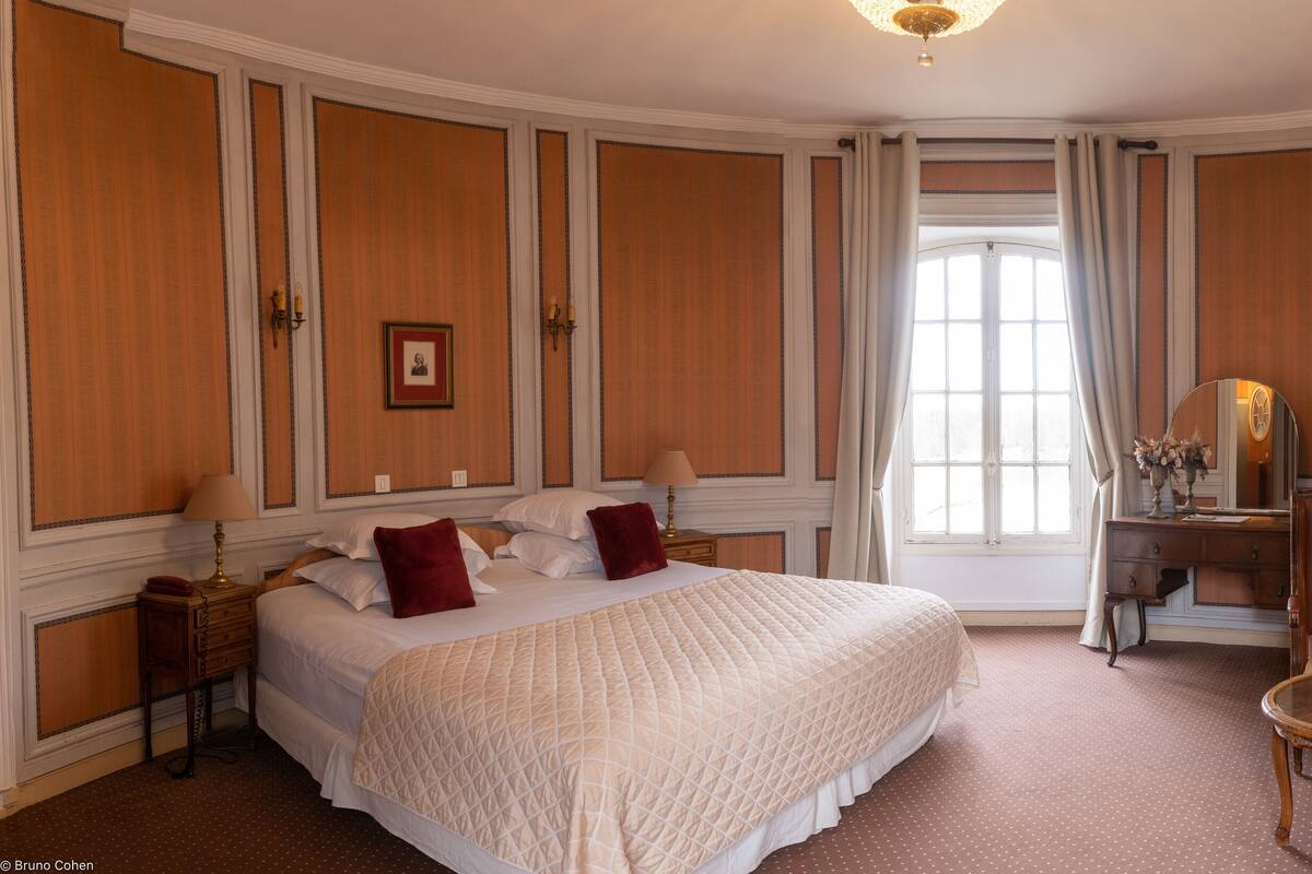 Une chambre d’hôtel avec un grand lit, un coin salon avec une table et des chaises, un miroir et une cheminée.