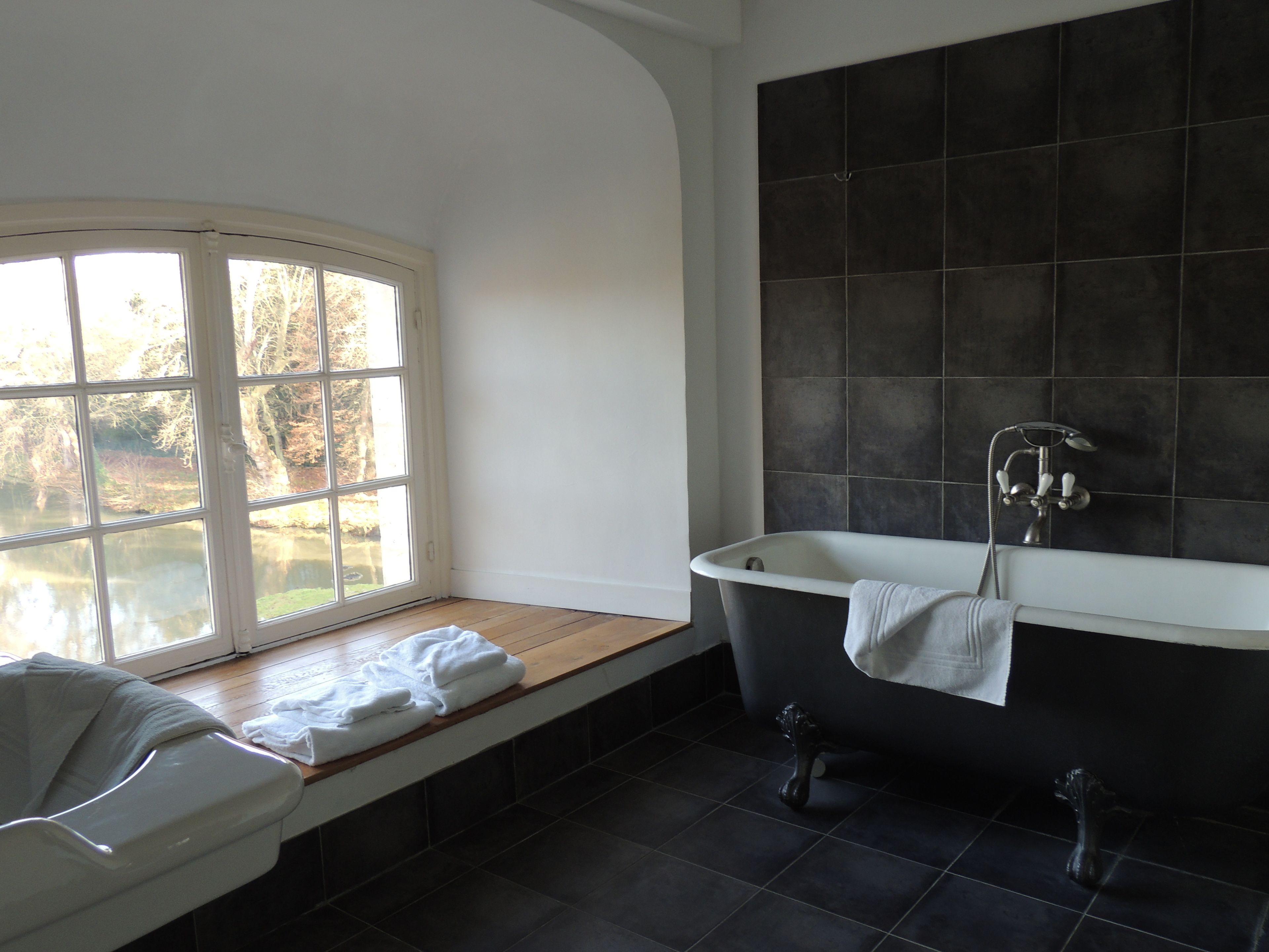 A bathroom with a bathtub, a window with a view of trees, and dark tiled walls.