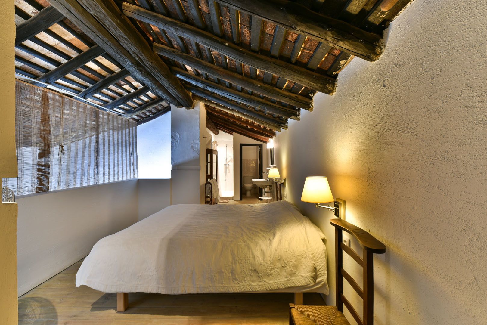 Un dormitorio con techo de madera, una cama, una mesita de noche, una ventana con vistas y un baño visible a través de una puerta abierta.