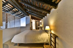 Un dormitorio con techo de madera, una cama, una mesita de noche, una ventana con vistas y un baño visible a través de una puerta abierta.
