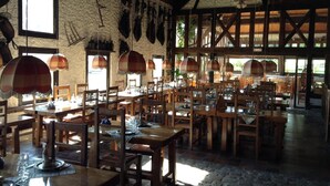 A rustic dining area with wooden tables and chairs, hanging lamps, and a stone wall with mounted items. - Hôtel Restaurant La Mangeoire – Relais Paris Bâle (Montieramey)