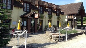 A traditional building with a 'BAR' sign, outdoor seating with umbrellas, and a stone fire pit. - Hôtel Restaurant La Mangeoire – Relais Paris Bâle (Montieramey)