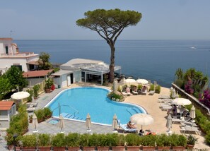 A pool area with a clear view of the sea, surrounded by lounge chairs and umbrellas, and a large tree nearby. - L'approdo (Casamicciola Terme)
