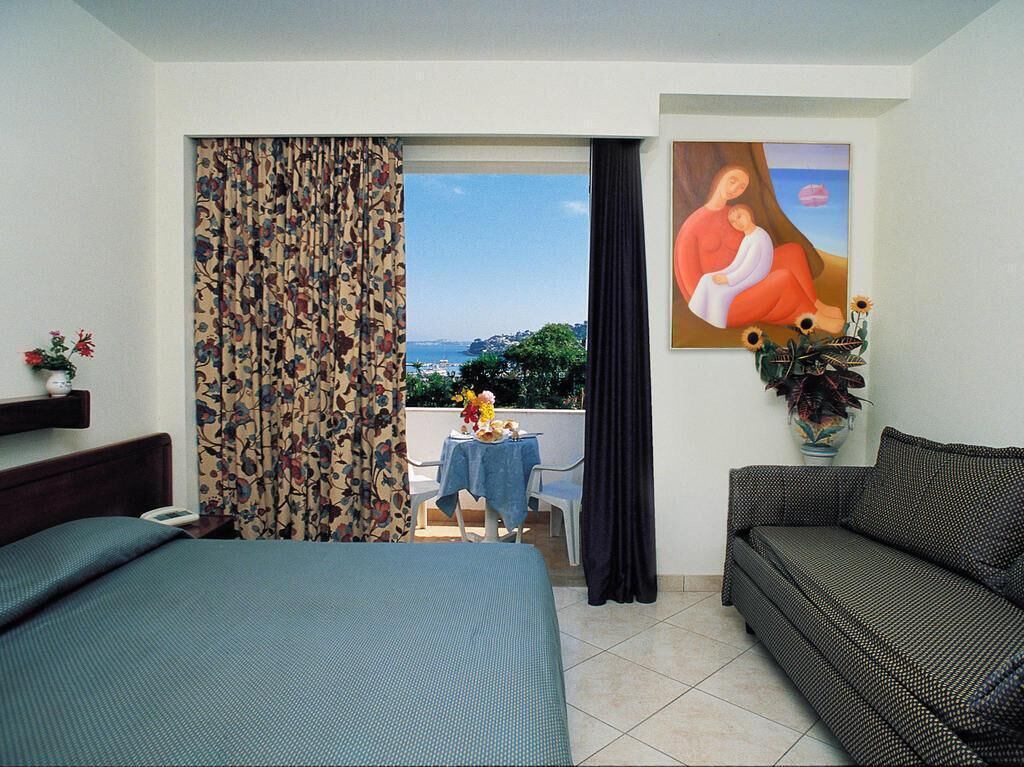 A hotel room with a bed, a sofa, a table with a vase of flowers, and a painting on the wall.