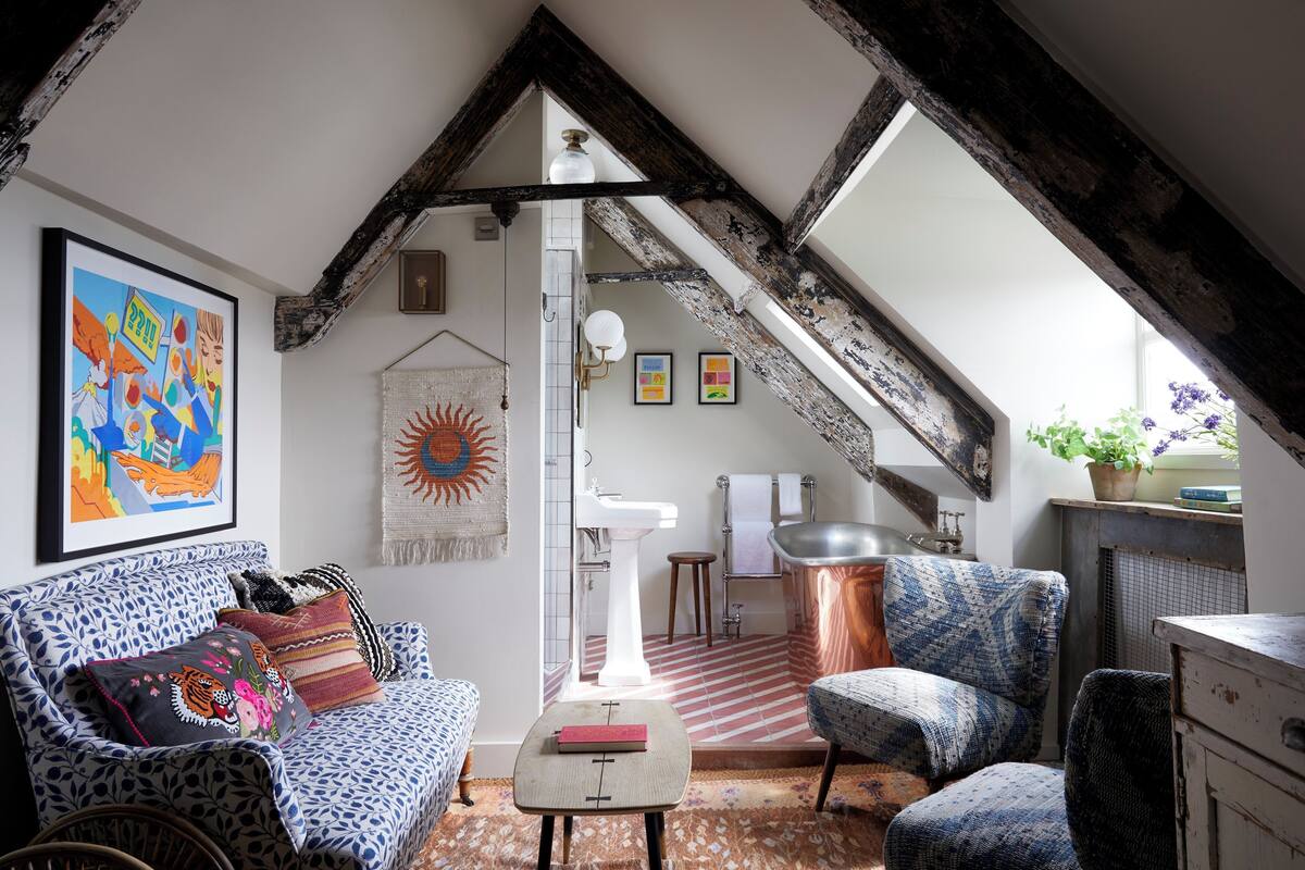 A cozy loft bedroom with exposed wooden beams, a large window, a white sink, a patterned sofa, and a small table.