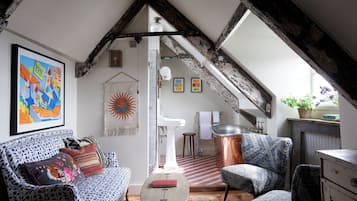 A cozy loft bedroom with exposed wooden beams, a large window, a white sink, a patterned sofa, and a small table.