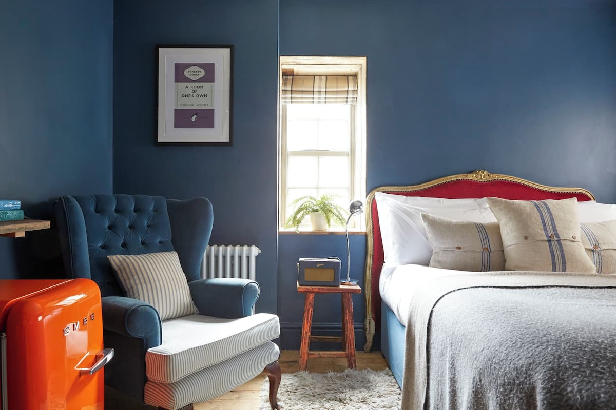 A bedroom with a blue wall, a tufted armchair, a vintage orange radio, a bed with a red headboard, and a framed picture on the wall.