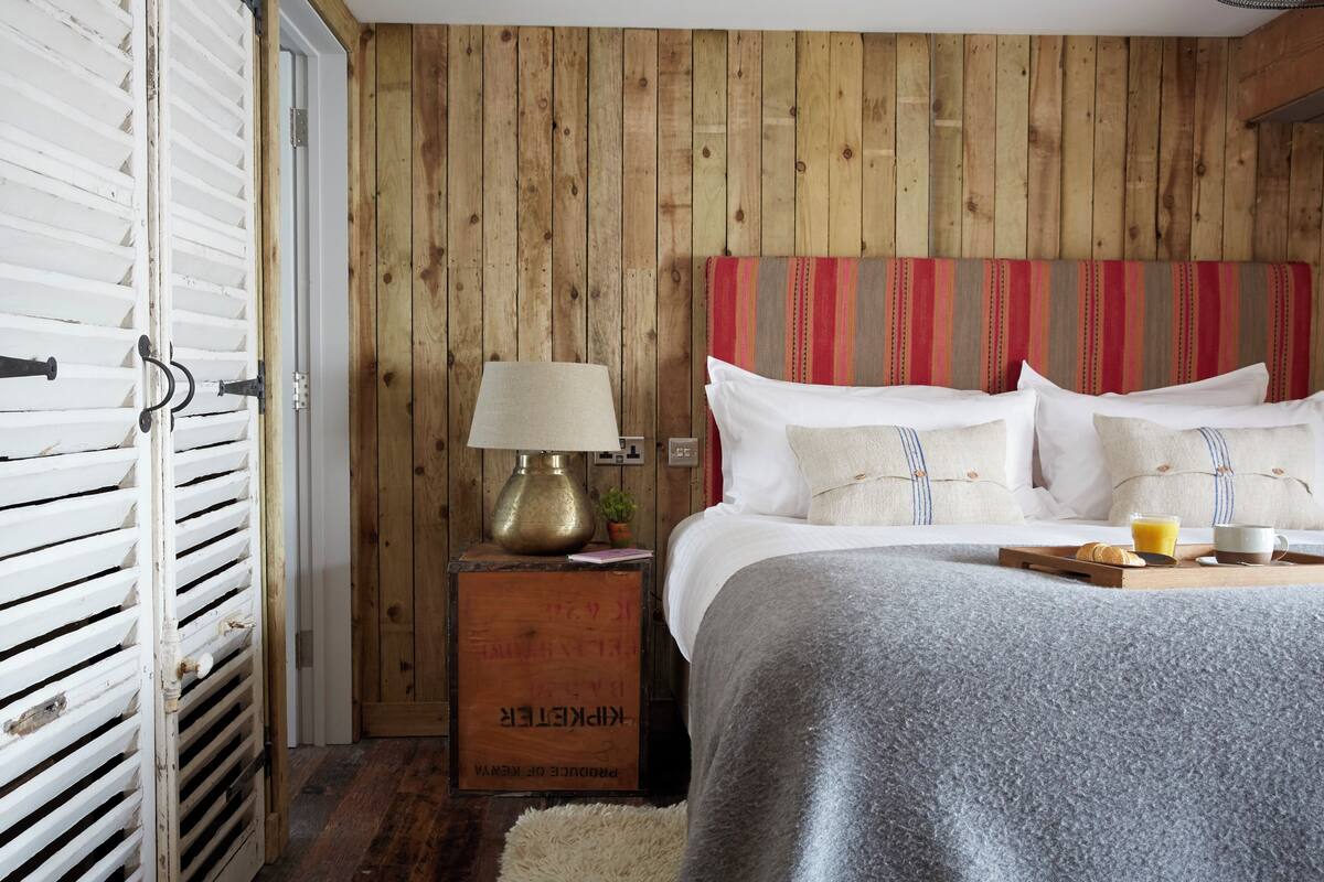 A bedroom with a wooden wall, a bed with white and striped bedding, a bedside lamp, a tray with breakfast items, and a vintage suitcase.
