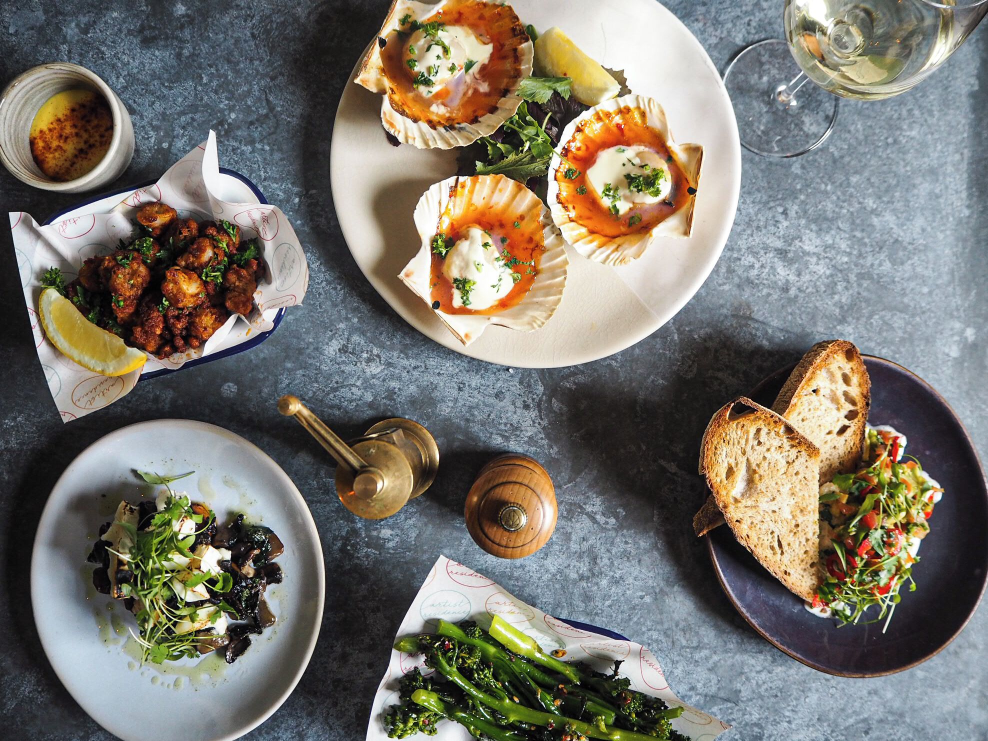 A variety of dishes including scallops, a salad, and bread, arranged on a table with a glass of wine.