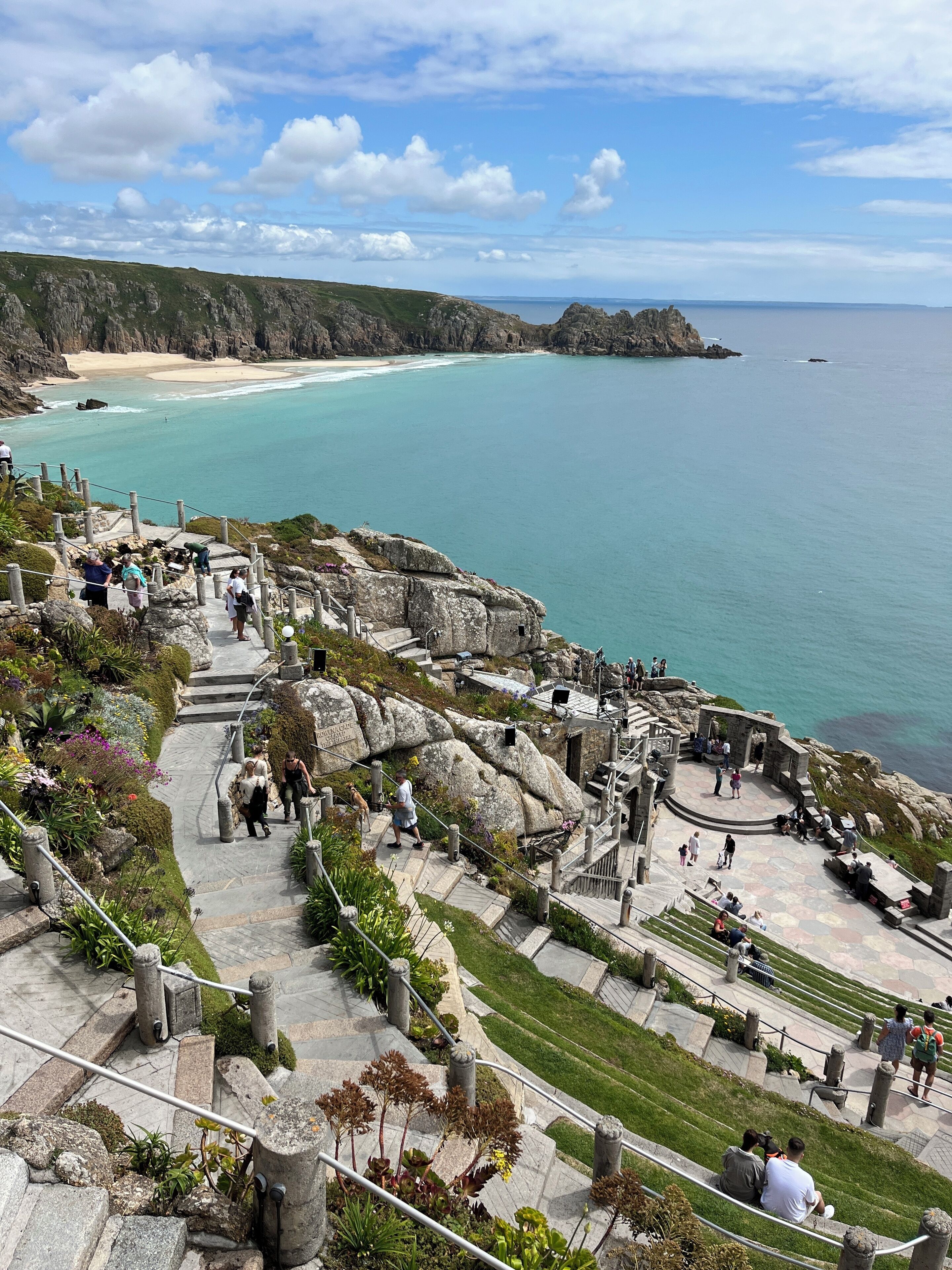 A coastal walkway with steps leading down to a beach, surrounded by rocky terrain and greenery.