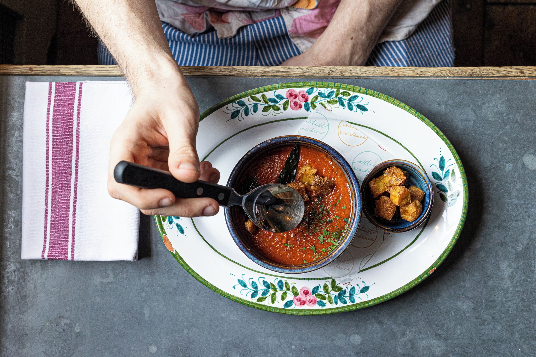 A person serving a bowl of soup with a spoon on a decorative plate.